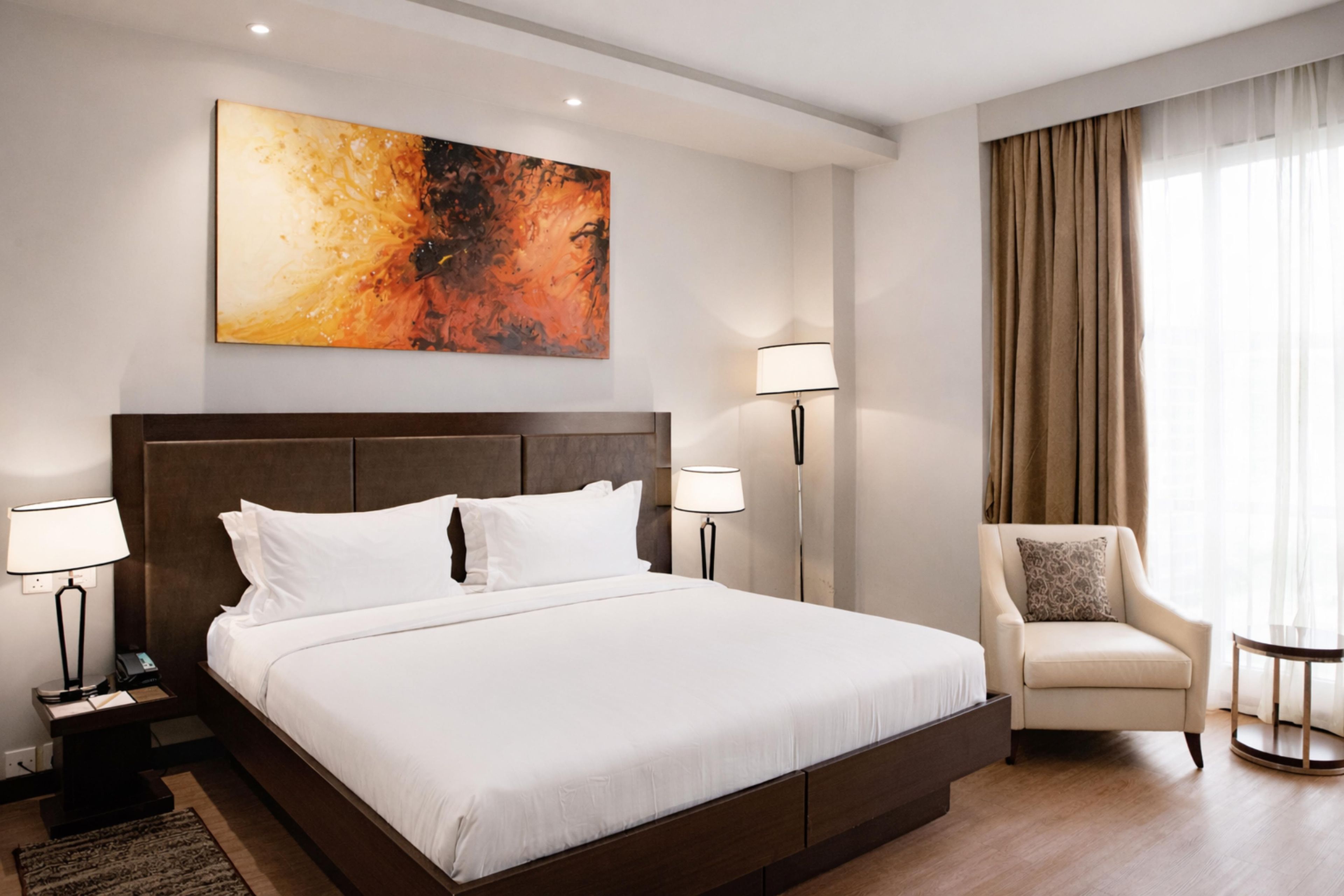 Habitación de hotel con una cama grande, dos mesitas de noche con lámparas, una silla y un cuadro en la pared.