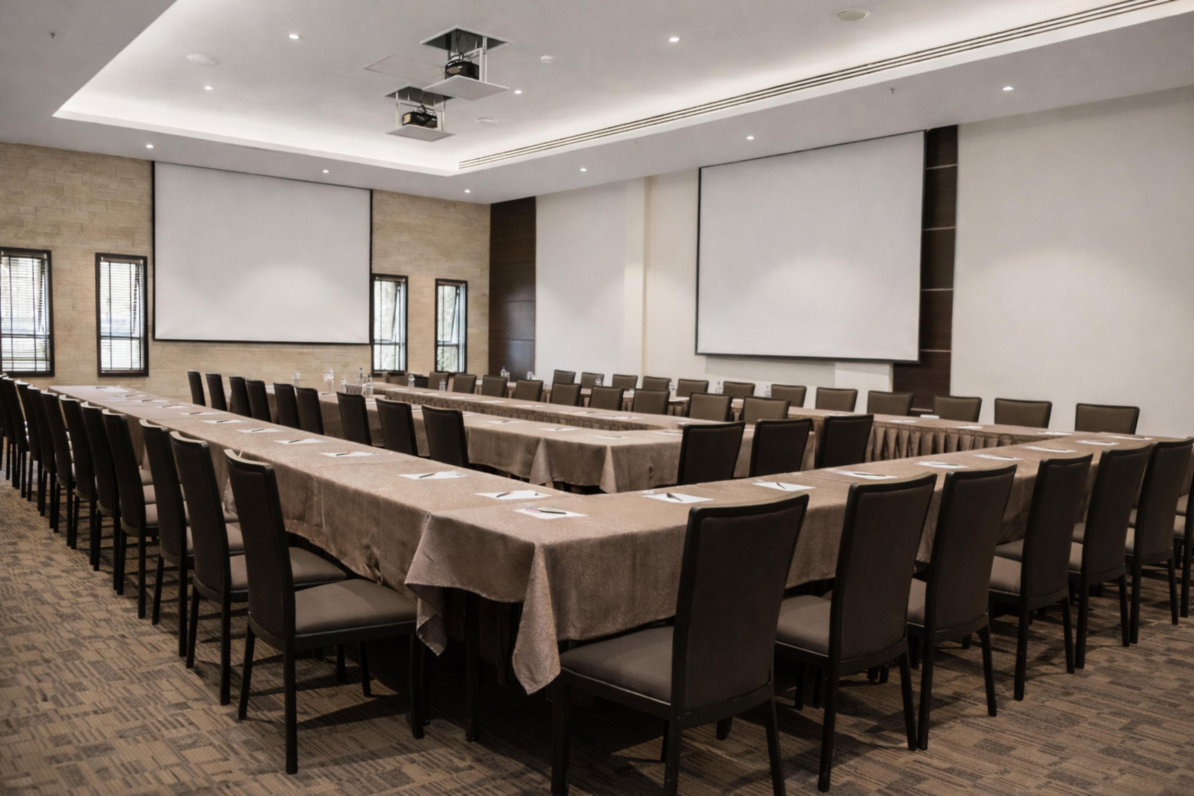 Une salle de conférence équipée de longues tables, de chaises, d’un projecteur et d’écrans.