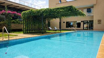 A swimming pool with a pergola covered in greenery, lounge chairs, and a building with a modern design in the background.