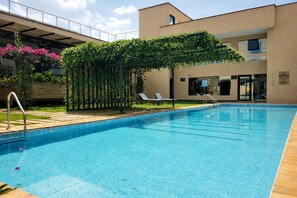 Una piscina con una pérgola cubierta de follaje, sillones de descanso y un edificio de diseño moderno al fondo.