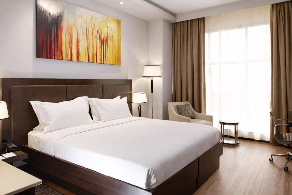 Ein Hotelzimmer mit einem großen Bett, einem Schreibtisch, einem Sessel, einer Lampe, einem Gemälde und einem Fenster mit Vorhängen.