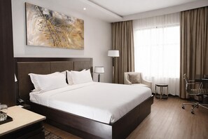 Habitación de hotel con una cama grande, un escritorio, una silla, una lámpara y un cuadro en la pared.
