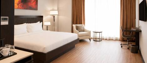 Une chambre d’hôtel avec un grand lit, un bureau avec une chaise, une petite table, une lampe, une télévision et un tableau au mur.
