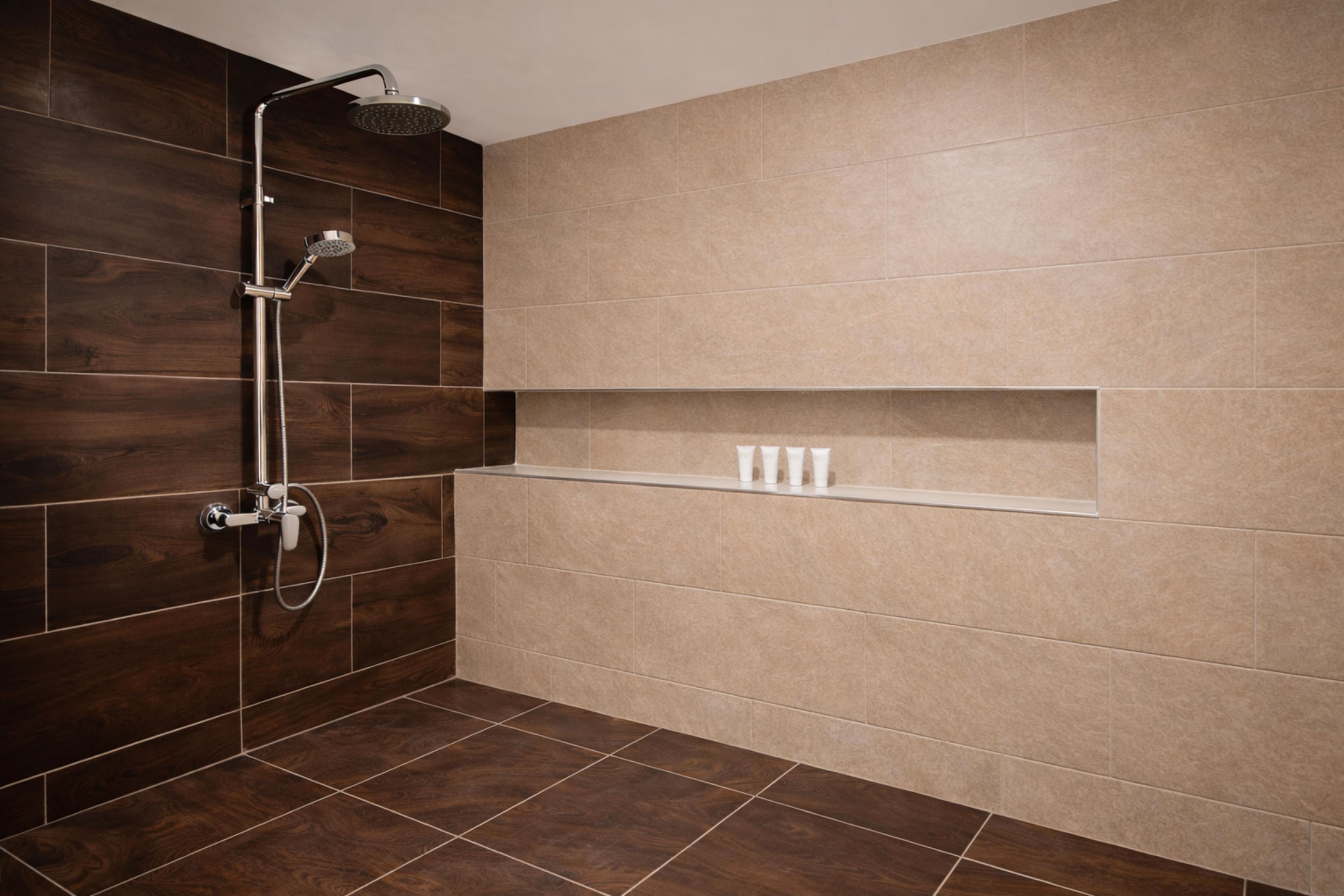 Un baño moderno con suelo de madera oscura, azulejos color beige, una ducha con cabezal de ducha manual y un estante con artículos de aseo.