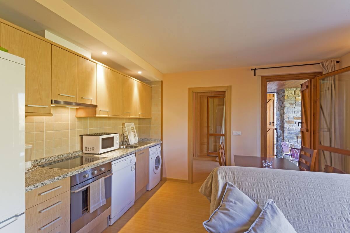 Cocina compacta con microondas, horno y lavadora. Posee una cama con manta y almohadas, y una zona de comedor con mesa y sillas.