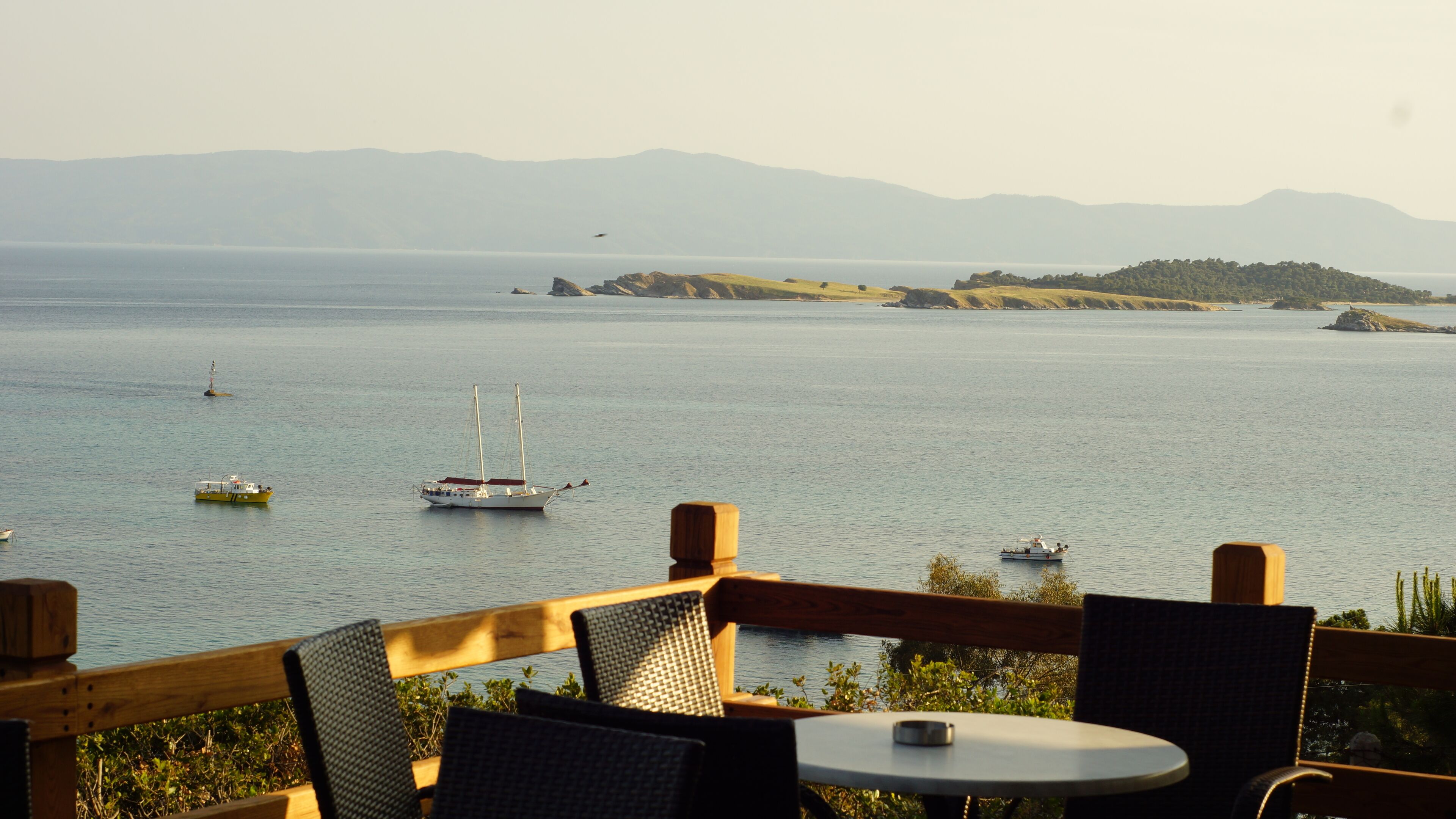 Un restaurante frente al mar con terraza y vistas a aguas tranquilas, barcos y colinas a lo lejos.