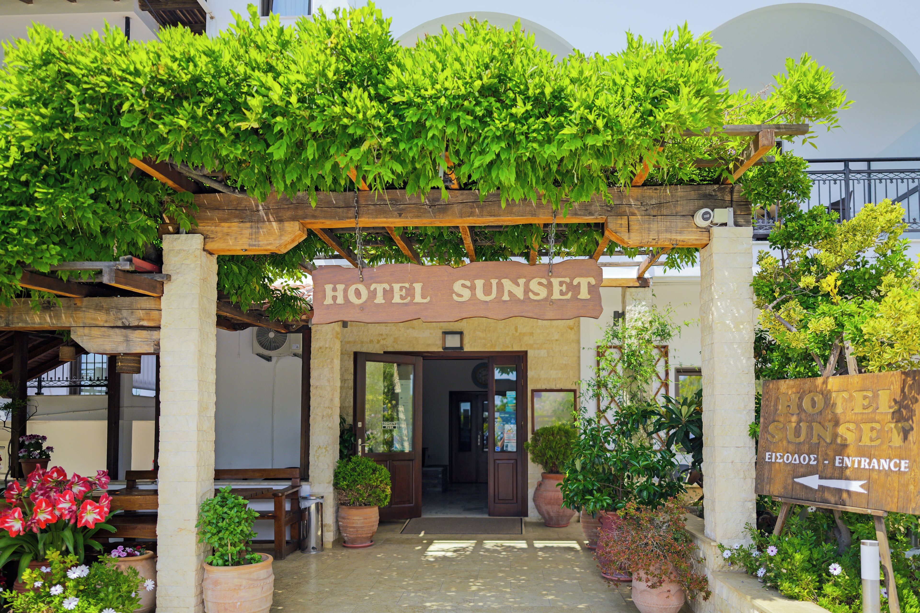 A hotel entrance with a wooden archway, a sign reading 'HOTEL SUNSET,' and potted plants around.