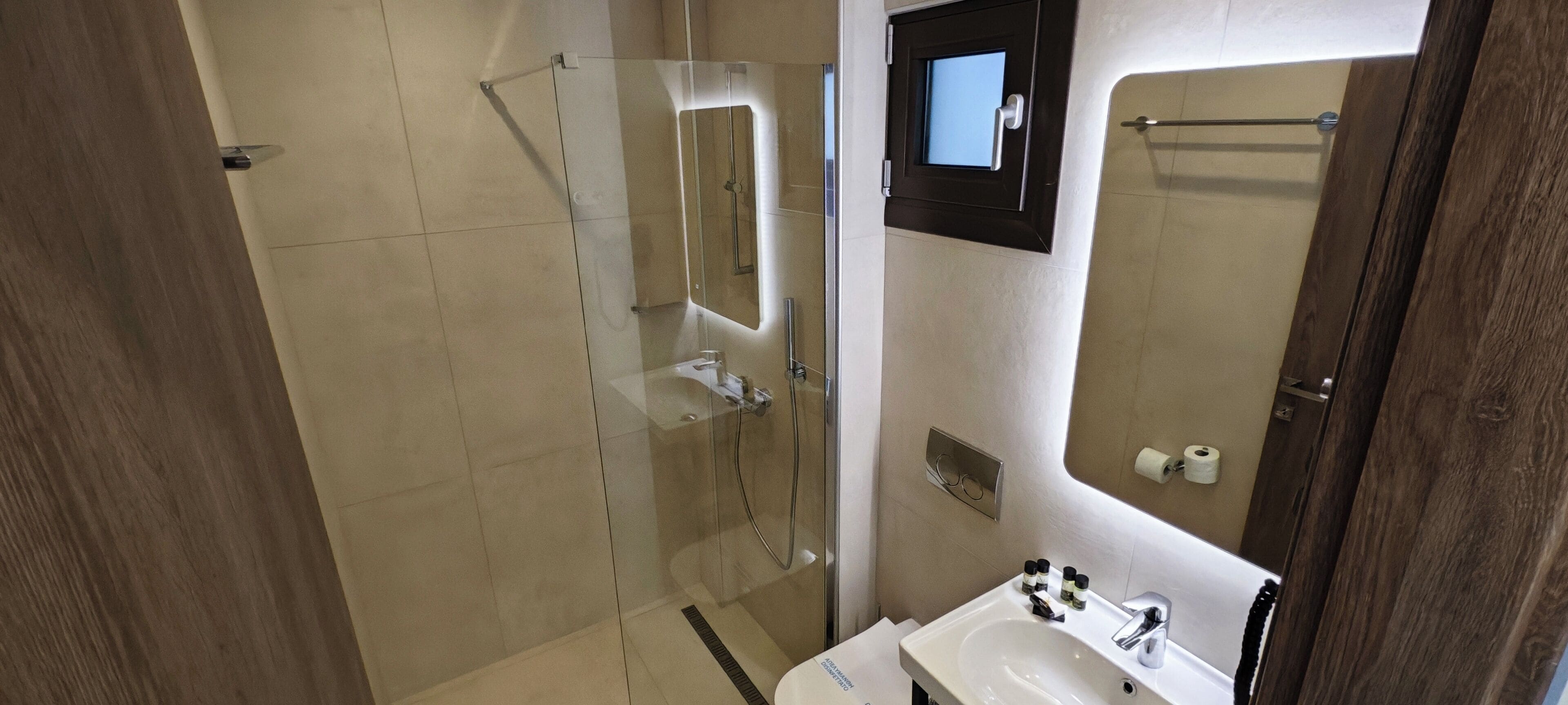 Une salle de bain moderne avec une douche à l’italienne en verre, un lavabo blanc et un miroir agrémenté d’une petite fenêtre.