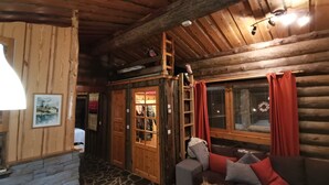 A cozy log cabin interior with a stone fireplace, a wooden ladder, a cushioned sofa, and a dining table with a bowl of fruit.