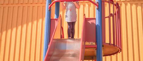 A child on a playground slide with a wooden building in the background.