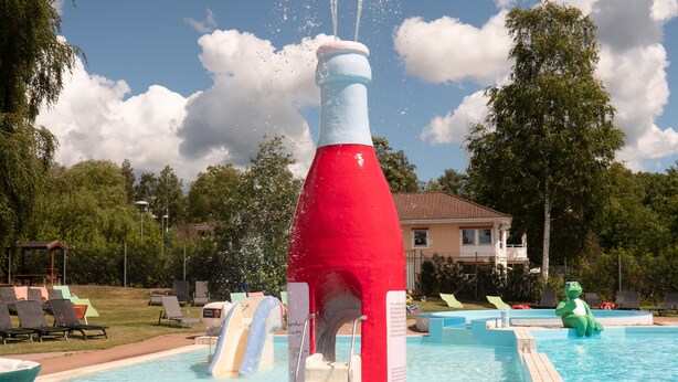 A swimming pool with a large red and white bottle-shaped fountain, slides, and lounge chairs. - Björkbackens Stugby - Campground (Vimmerby)