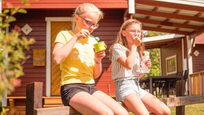 Two people sitting on a wooden bench outside a red building with a white window and a small porch area. - Björkbackens Stugby - Campground (Vimmerby)