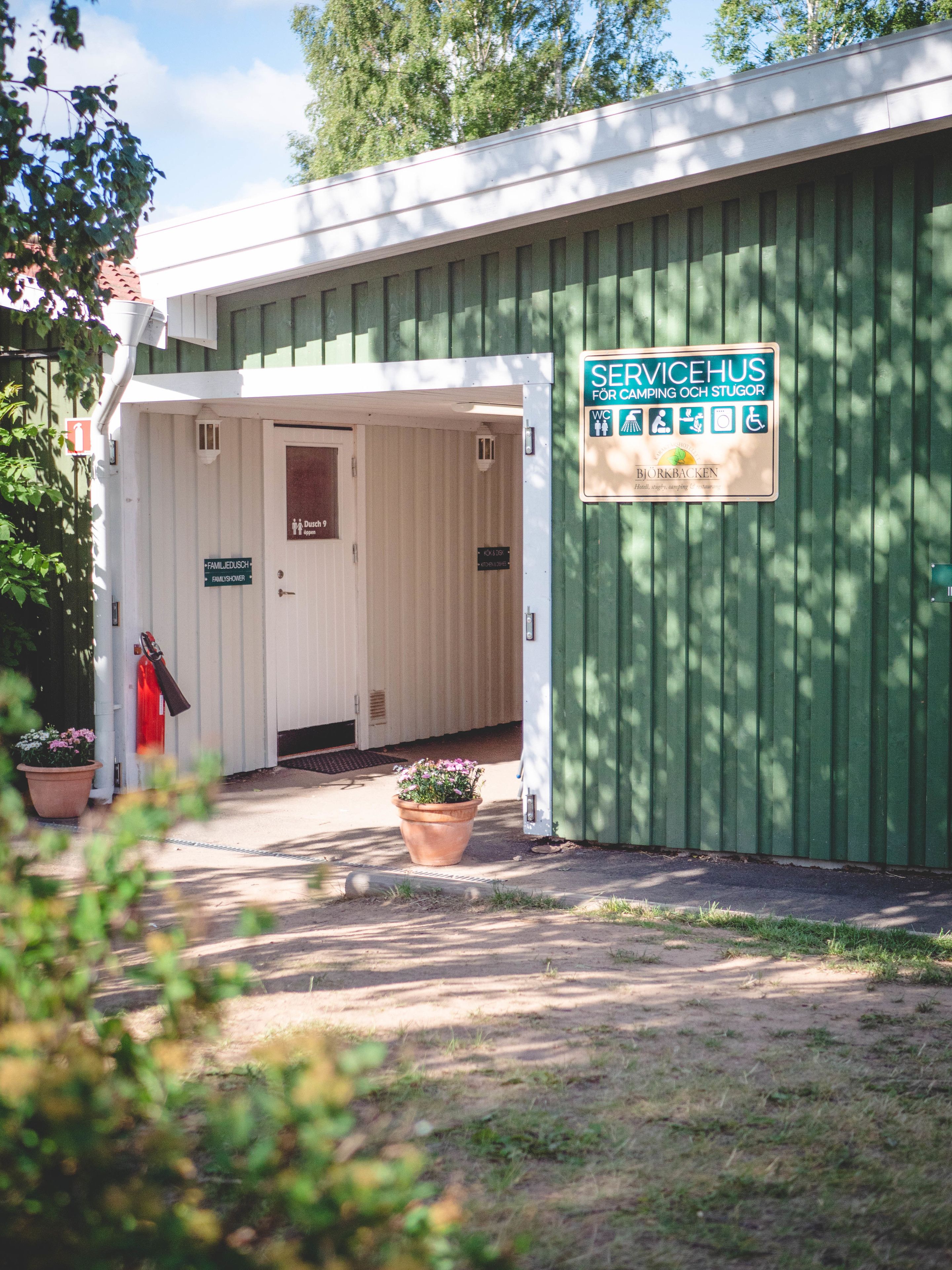 Un edificio verde con un letrero que indica que es una casa de servicios para camping y establos. Se ve una puerta con la leyenda "FAMILIEDUSCH" y un cartel para ducha familiar.