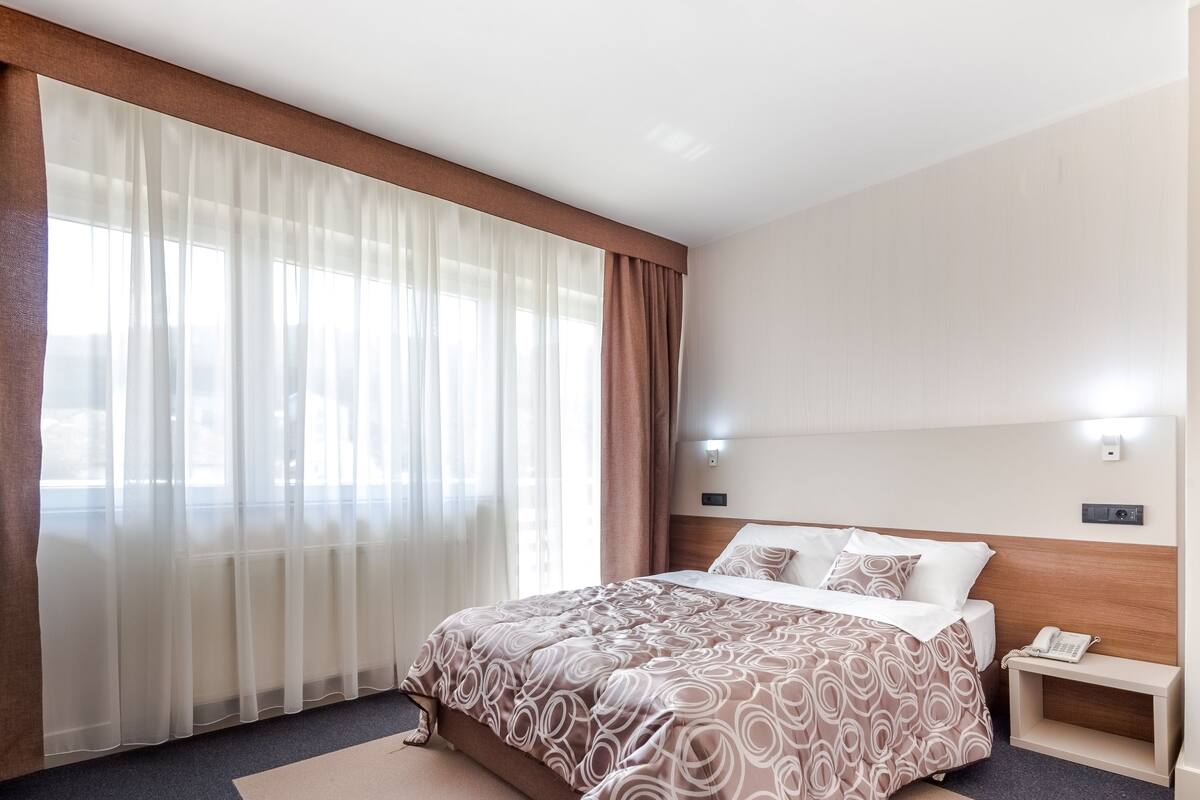 A hotel room with a large bed, a bedside table with a phone, and a window with sheer curtains.