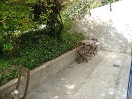 Un patio con una mesa y sillas de hormigón, rodeado de follaje denso y una pared blanca.