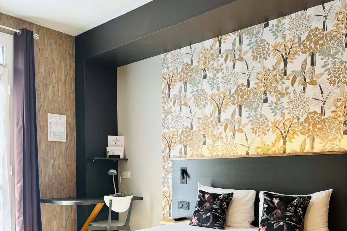 A modern hotel room with a large bed, a headboard with a floral pattern, a desk with a chair, and a window with curtains.
