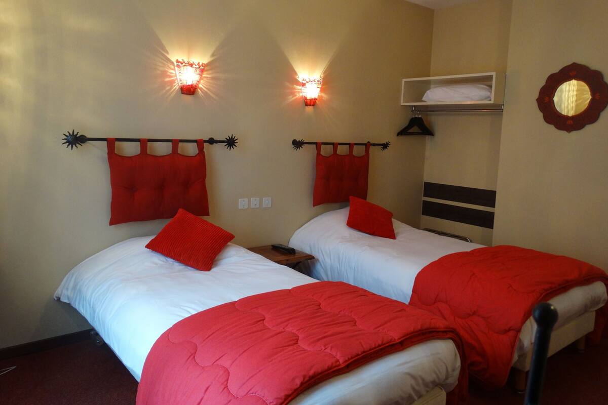 A hotel room with two beds, red bedding, wall-mounted lights, a mirror, and a shelf with a towel.