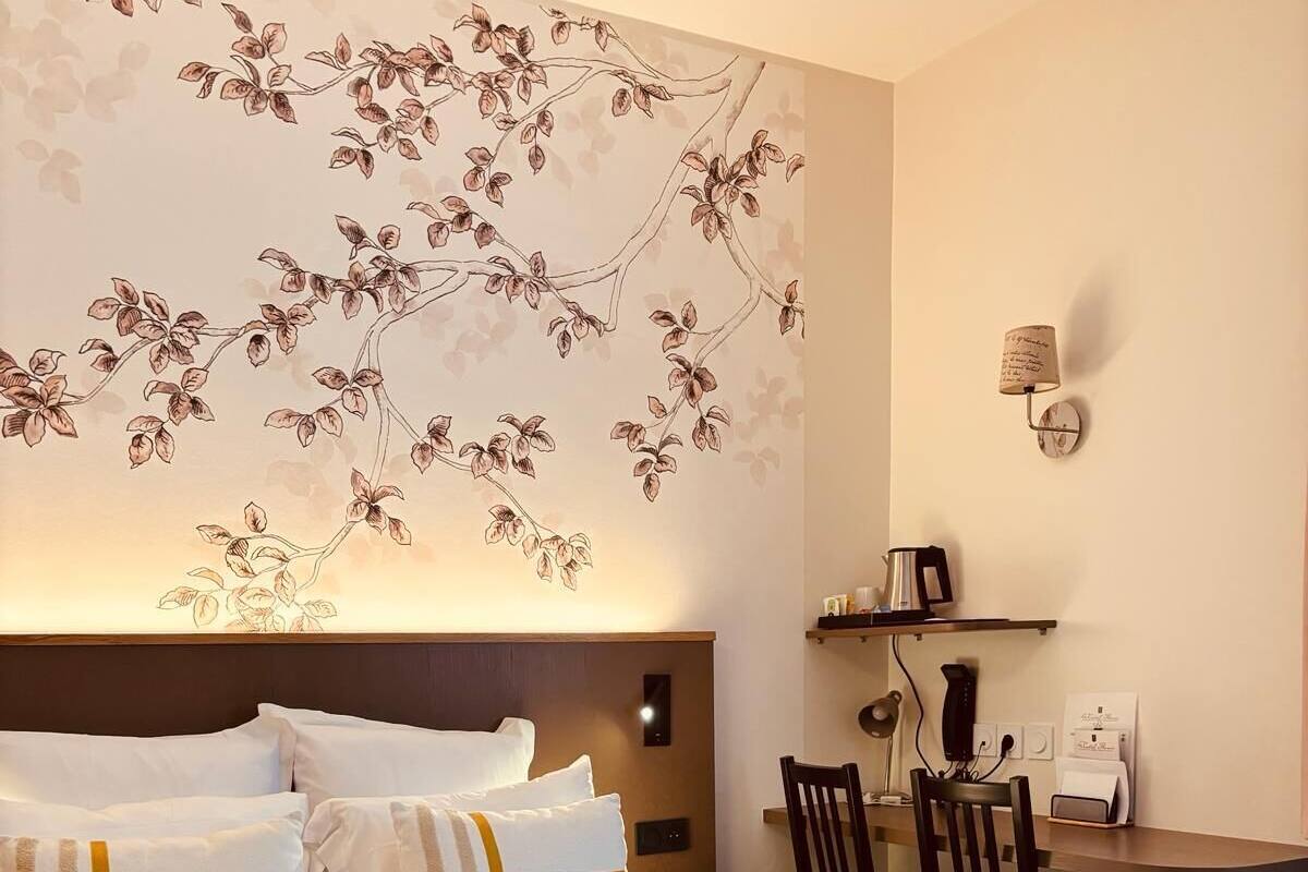 A hotel room with a large bed, a desk with a chair, a coffee maker, and a wall-mounted lamp.