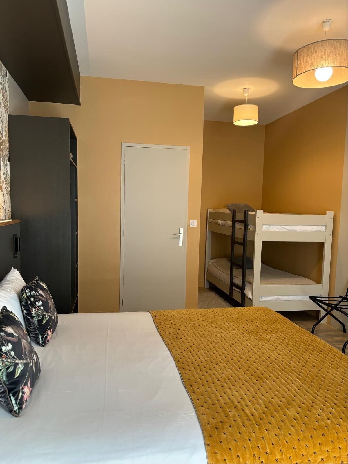 A bedroom with a bunk bed, a bed with a yellow blanket, a wardrobe, and a door.
