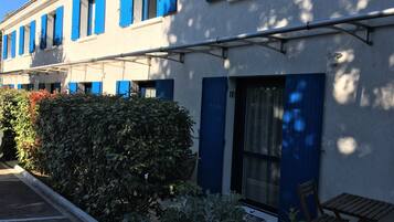 Un bâtiment avec des volets bleus, une plaque portant le numéro 18, et un petit espace extérieur aménagé avec une chaise et une table.