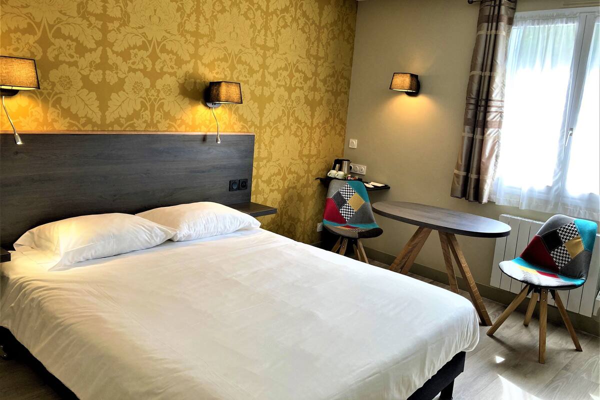 A hotel room with a bed, two wall-mounted lamps, a small table, a chair, and a window with curtains.