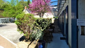 A covered outdoor seating area with tables and chairs, a building entrance with a sign, and a tree with purple flowers. - Hotel Le Castel Fleuri (Tours)