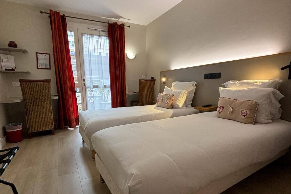 A hotel room with two beds, a wicker chair, a small table, and a view of the outside through a window with red curtains.