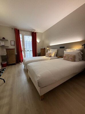A hotel room with two beds, a wicker chair, a small table, and a view of the outside through a window with red curtains. - Hotel Le Castel Fleuri (Tours)