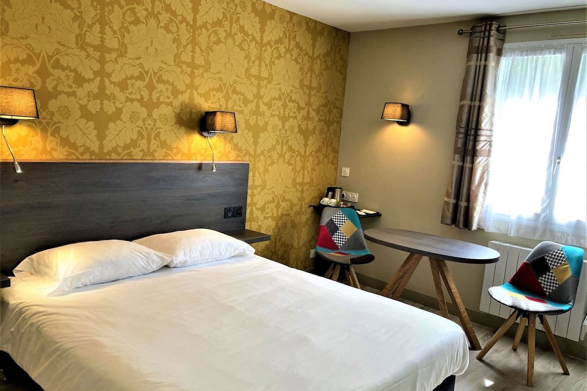 A hotel room with a bed, two wall-mounted lamps, a small table, a chair, and a window with curtains.