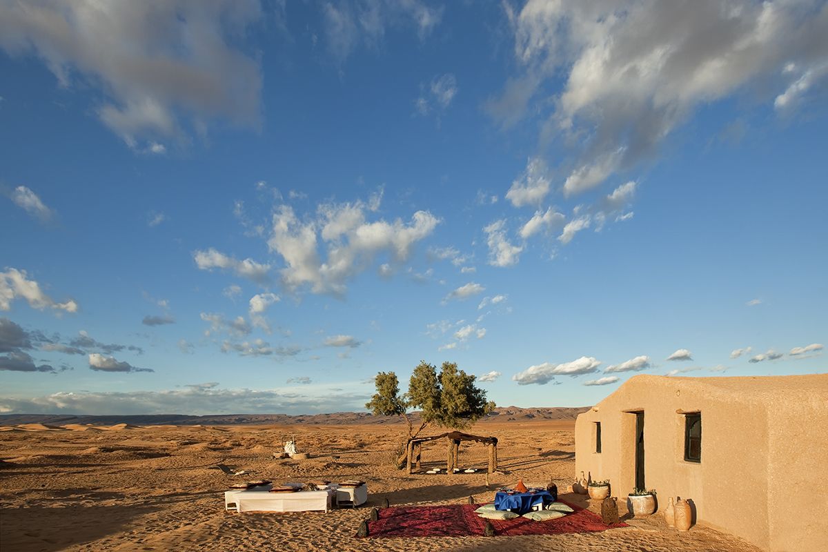 A desert landscape with a small building, a tree, and a tent.