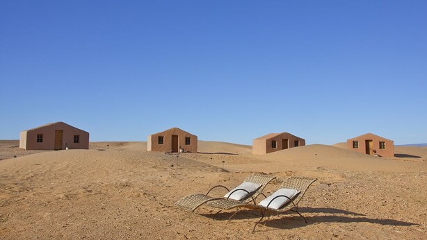 A desert landscape with four small, single-story buildings and two sun loungers in the foreground. - La Dune Blanche (M'Hamid El Ghizlane)