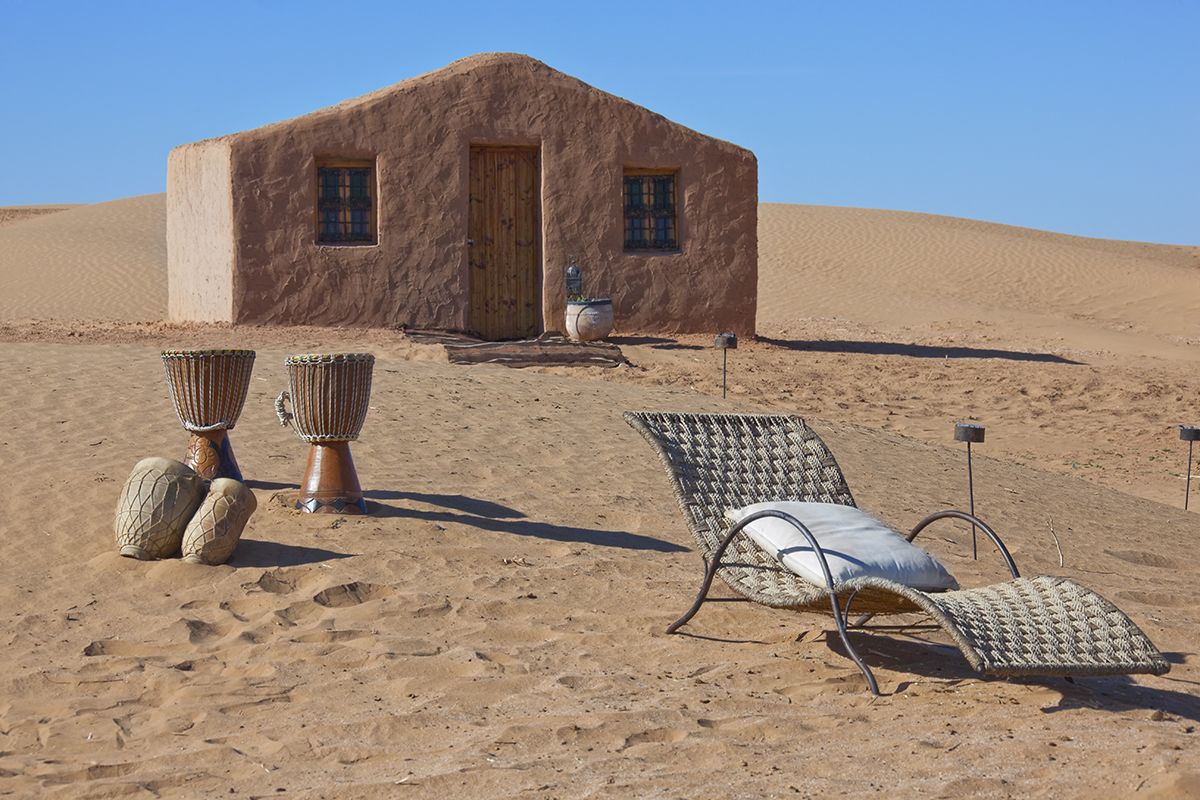 A desert scene with a single-story adobe building, two large woven chairs, and a single lounge chair with a cushion.