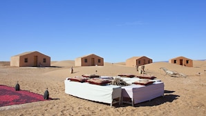 A desert setting with a dining area set up on the sand, featuring a table with pillows and a red carpet. - La Dune Blanche (M'Hamid El Ghizlane)