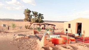 A desert setting with a small building, outdoor seating, and a tree. - La Dune Blanche (M'Hamid El Ghizlane)