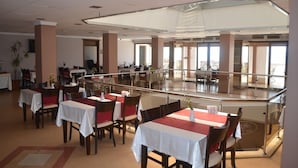 A spacious dining area with multiple tables and chairs, a glass railing, and large windows allowing natural light. - Ayvalik Palas Hotel (Ayvalik)