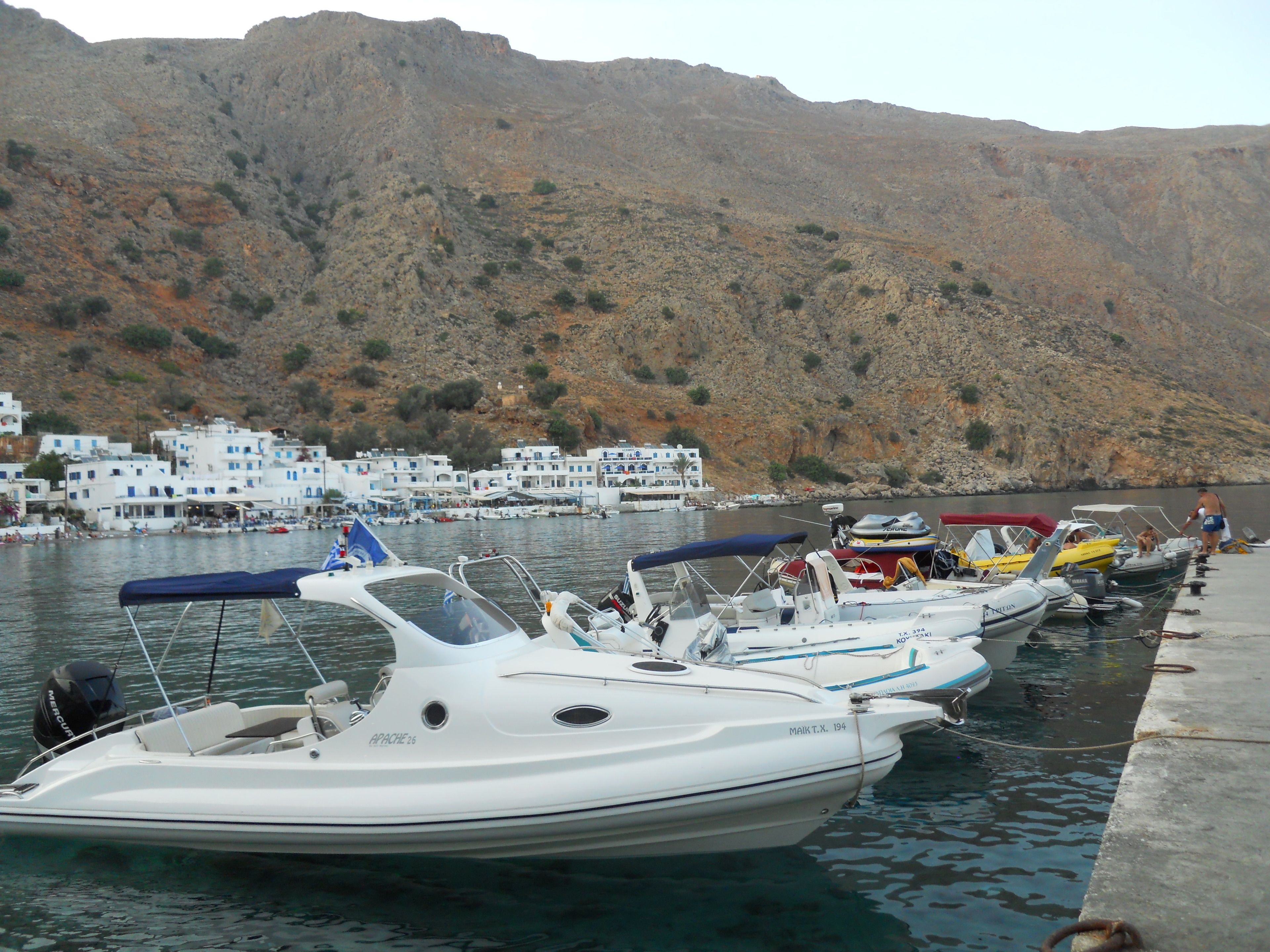 Un puerto deportivo con varias embarcaciones amarradas, una ladera rocosa al fondo y edificios blancos con detalles azules.