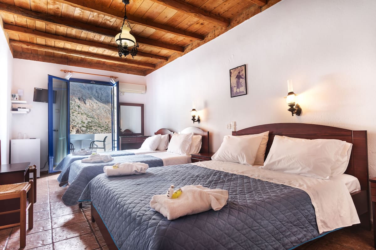 A hotel room with two beds, wooden ceiling beams, a balcony with a mountain view, and a small table with chairs.