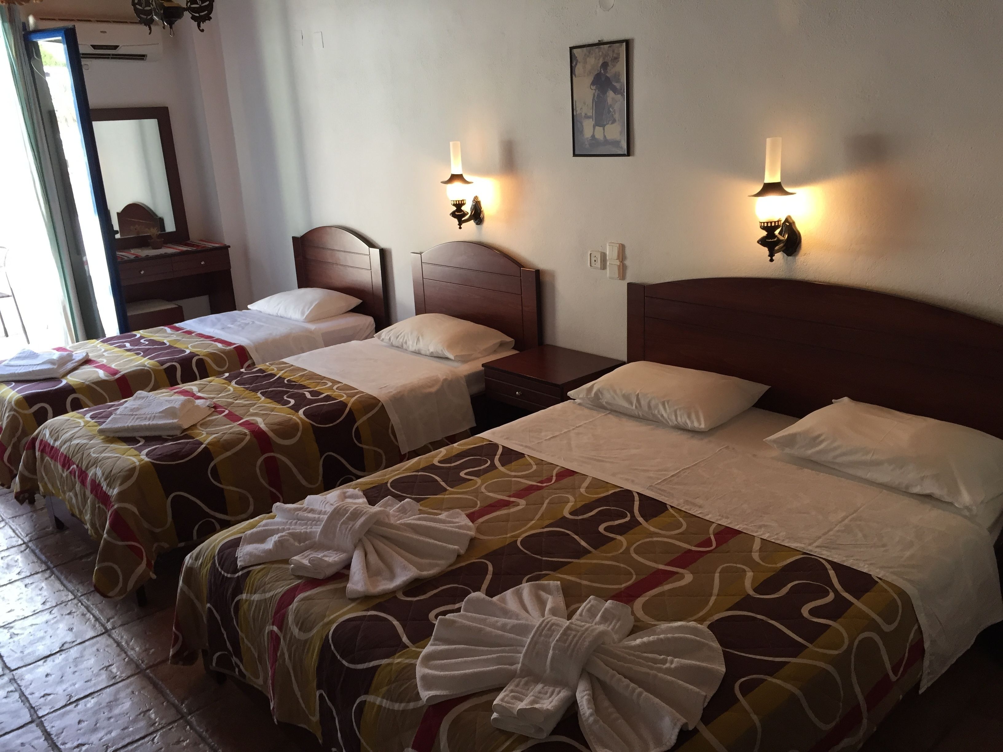 A hotel room with two beds, each with a patterned bedspread and a towel. There are wall-mounted lights and a framed picture on the wall.