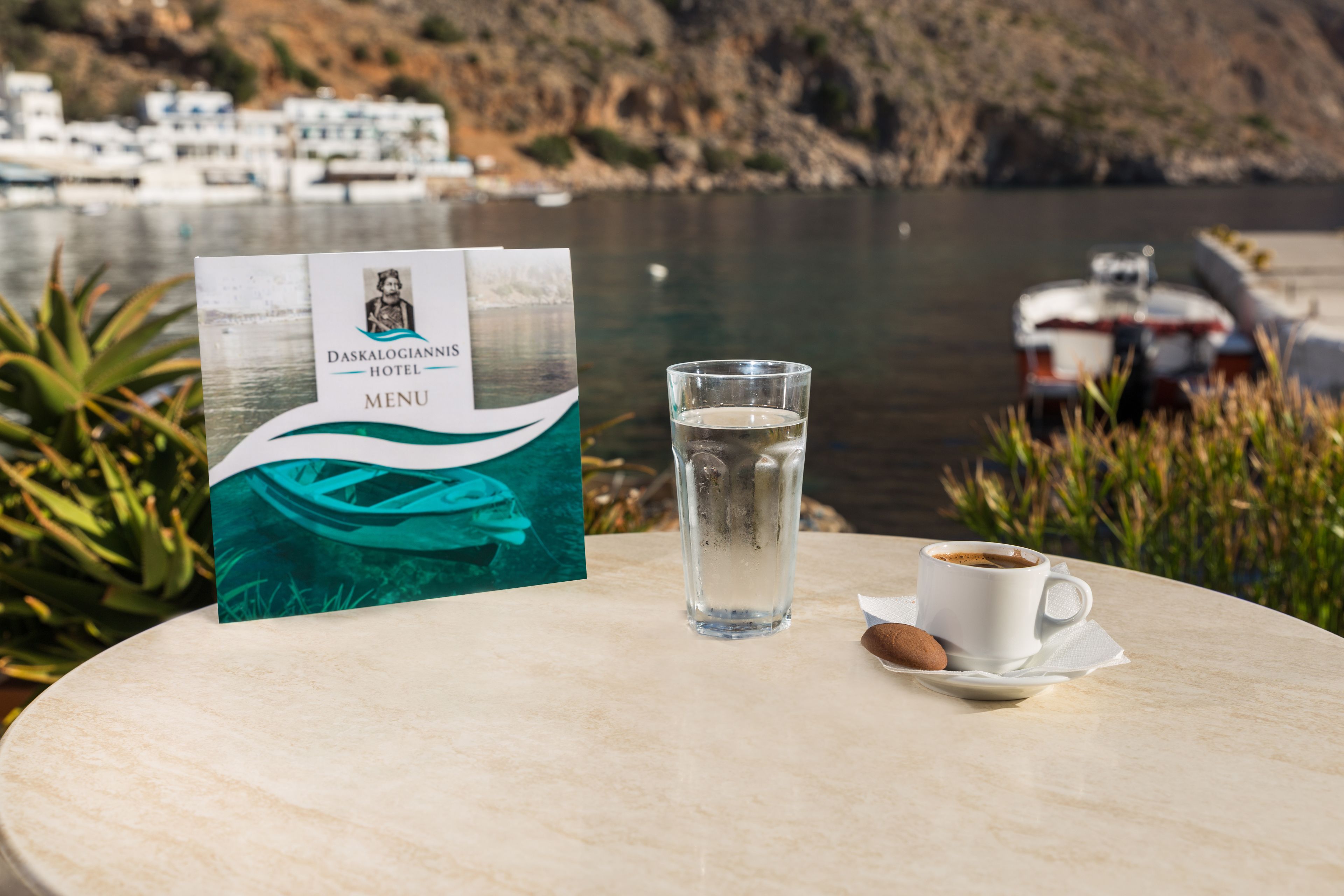 Speisekarte vom Daskalogiannis Hotel auf einem Tisch mit einem Glas Wasser und einer Tasse Kaffee, mit Blick auf eine Küstenlandschaft mit Booten und Gebäuden.