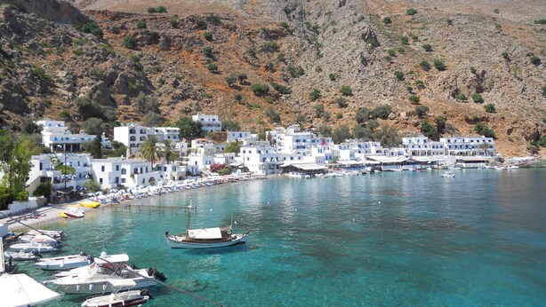 A coastal town with white buildings, a marina with boats, and a mountainous backdrop. - Daskalogiannis Hotel (Sfakia)