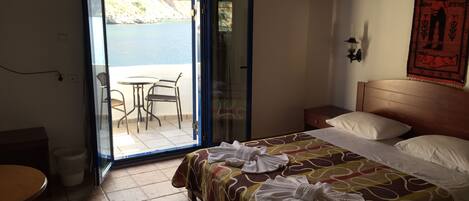 A hotel room with a bed, a small table, and a chair near a sliding glass door leading to a balcony with a view of the sea.