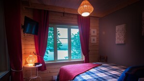 Un dormitorio con cama, televisor montado en la pared, ventana con cortinas, mesita de noche con lámpara y un cuadro en la pared.