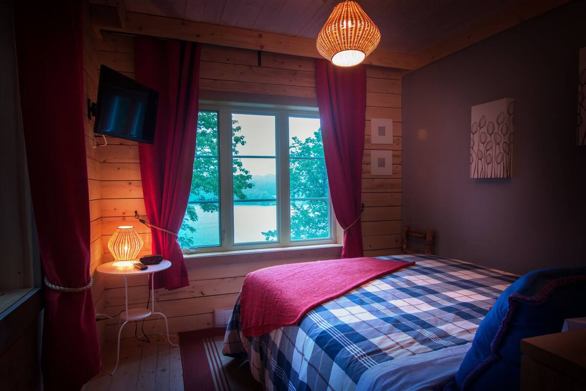 A bedroom with a bed, a television mounted on the wall, a window with curtains, a table with a lamp, and a painting on the wall.