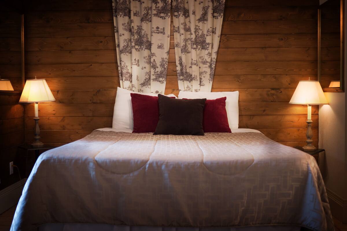 A neatly made bed with a white and beige patterned bedspread, two red and one dark brown pillow, and two bedside lamps on wooden nightstands.