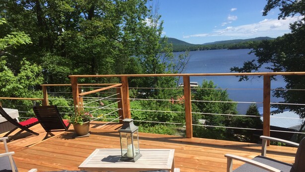Una terraza de madera con mobiliario exterior, una mesa de vidrio y vistas a un lago rodeado de árboles.