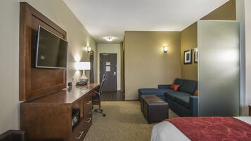 Quarto de hotel com cama, secretária com cadeira, sofá, televisão presa na parede e porta que dá para outro espaço.