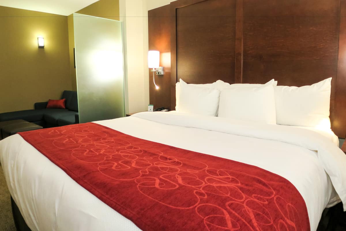 A hotel room with a large bed, a red patterned bedspread, a wooden headboard, a nightstand with a lamp, and a seating area with a couch and pillows.