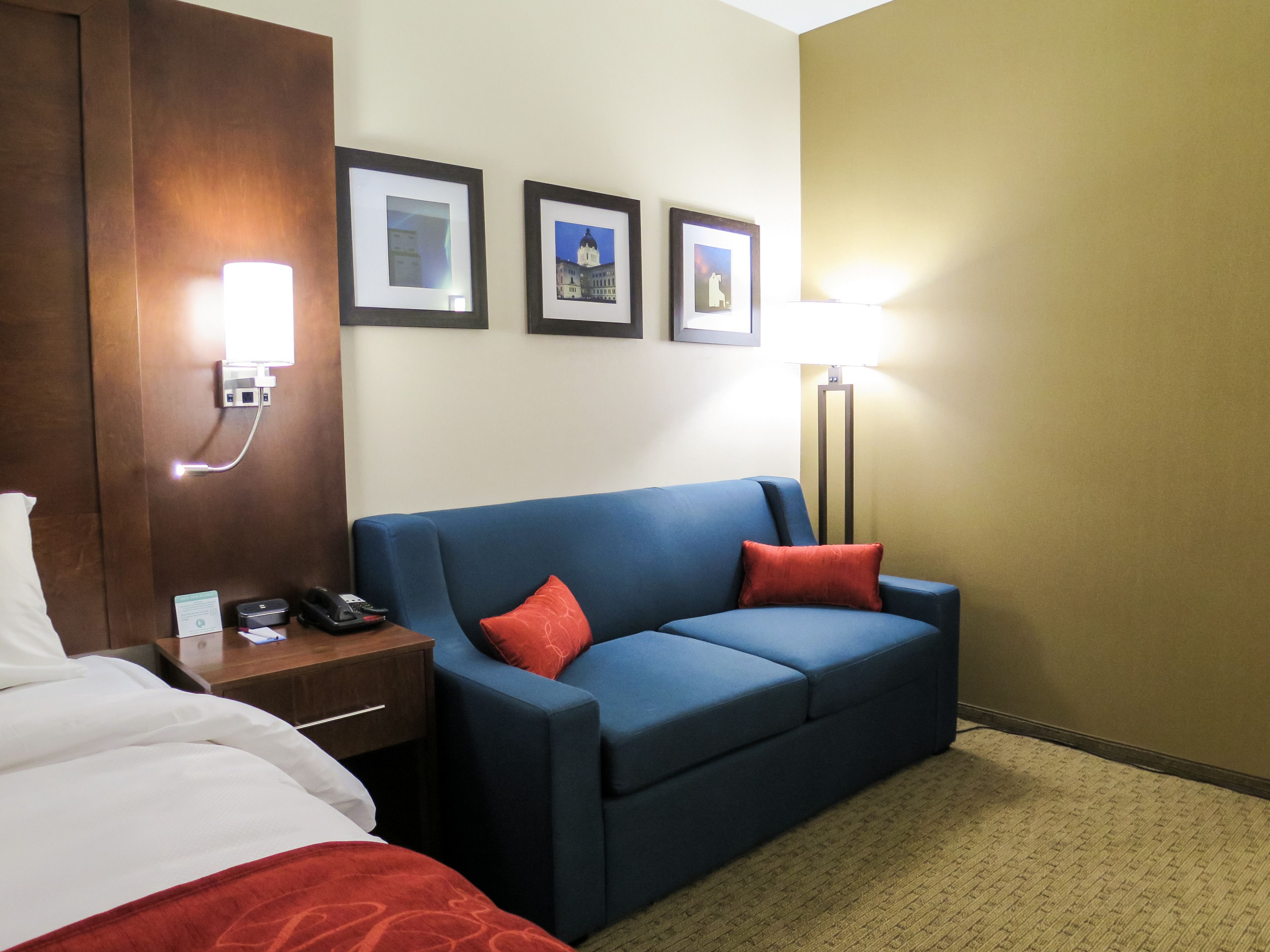 A hotel room with a bed, a blue sofa with red cushions, a bedside table with a phone, and framed pictures on the wall.