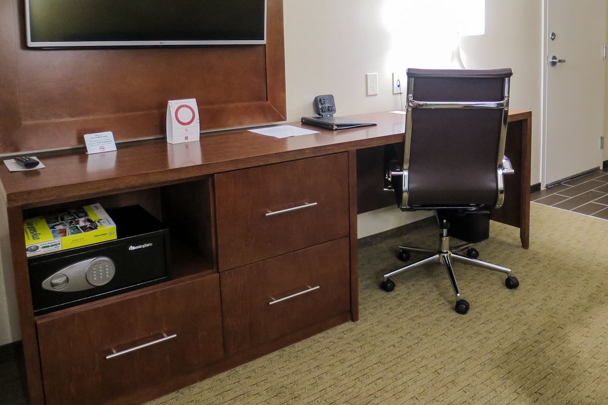 A hotel room with a wooden desk, a black office chair, a flat-screen TV mounted on the wall, a clock, and a small shelf with a box.