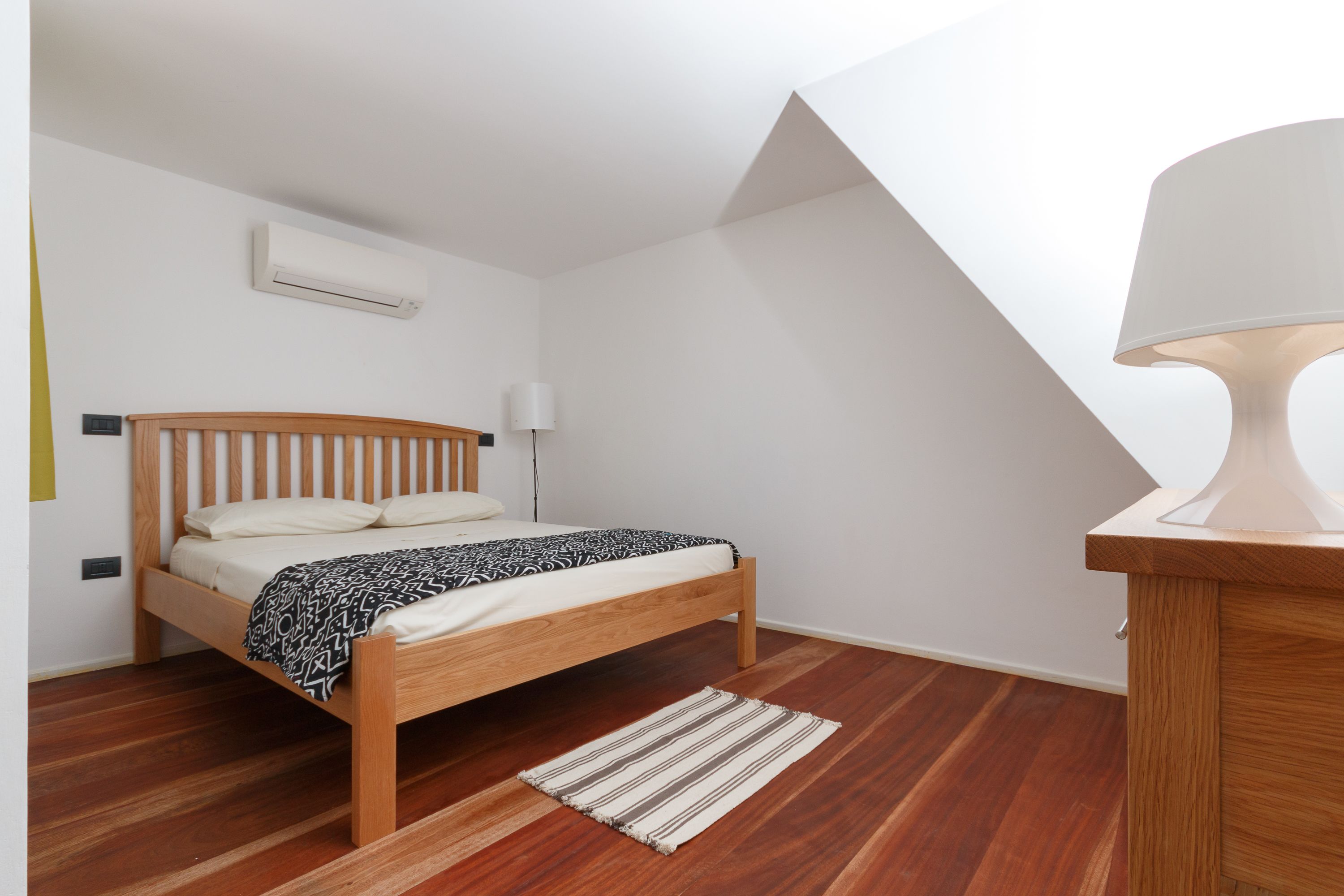A bedroom with a wooden bed, a white air conditioner, a bedside lamp, and a wooden nightstand.
